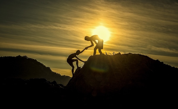 Person helping another climb a steep rocky hill at sunset, symbolizing teamwork and support